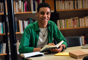 Wall Mural - Portrait of young male student in the library holding a book in his hands and looking at camera.