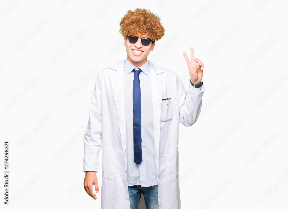 Young handsome scientist man wearing professional coat and sunglasses smiling with happy face winking at the camera doing victory sign. Number two.