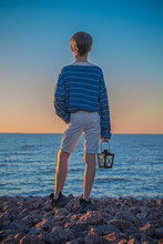 A Boy Stands With His Back And Looks At The Sea And The Sunset Sky, Rocky Beach, Old Black Lamp In His Hand