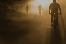 People Mountain Biking Downhill In Dust Backlit Silhouette