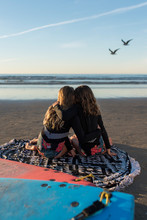 Twin Sisters Hugging While Sitting On The Beach Watching The Surf