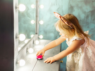 Little girl plays with toy pink car. Cute happy 4 year old baby girl . Modern nursery interior, early learning concept. Awesome daughter in white dress with mouse ears instead mirror. Selective focus