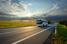 Two White Buses Traveling On The Asphalt Road In Rural Landscape At Sunset With Dramatic Clouds