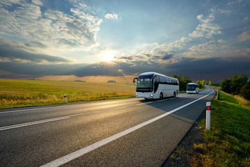 two white buses traveling on the asphalt road in rural landscape at sunset with dramatic clouds