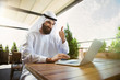 © master1305 - Arab saudi businessman working online with a laptop and tablet in a coffee shop or a cafe with an outdoor terrace in the background. Concept of business, finance, modern technologies, start up.