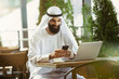 © master1305 - Arab saudi businessman working online with a laptop and tablet in a coffee shop or a cafe with an outdoor terrace in the background. Concept of business, finance, modern technologies, start up.