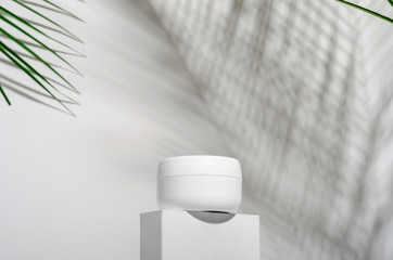 White jar of cream on a stand on a white background with tropical palm leaves and their shadow. Stylish look of the product, mock up, identity.