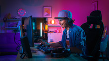 Professional Gamer Playing First-Person Shooter Online Video Game On His Powerful Personal Computer With Colorful Neon Led Lights. Young Man Is Wearing A Cap. Living Room Lit With Pink Lamps. Evening.