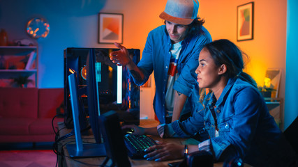 Wall Mural - Two Friends or a Couple Discuss Future Strategies on a Computer Game. Pretty Black Girl Shows Her Work to a Young Man and He Shares Creative Ideas. Cozy Room is Lit with Warm and Neon Light.