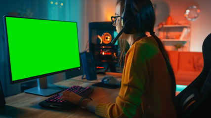 Wall Mural - Excited and Concentrated Gamer Girl in Glasses and Headset with a Mic Playing on Her Personal Computer with Green Screen Mock-up Template. Colorful Warm Neon Led Lights. Cozy Evening at Home.