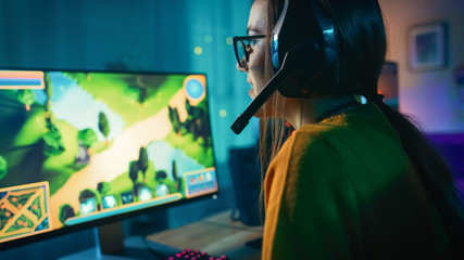 Wall Mural - Excited and Concentrated Gamer Girl in Glasses and Headset with a Mic Playing Online Strategy Video Game on Her Personal Computer. Room and PC have Colorful Warm Neon Led Lights. Cozy Evening at Home.