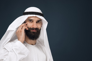 Half-length portrait of arabian saudi man on dark blue studio background. Young male model using smartphone, making a call. Concept of business, finance, facial expression, human emotions