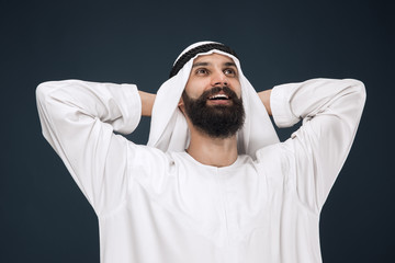 Half-length portrait of arabian saudi businessman on dark blue studio background. Young male model standing and resting. Concept of business, finance, facial expression, human emotions.