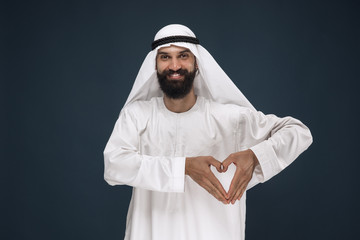 Portrait of arabian saudi businessman on dark blue studio background. Young male model standing a showing a gesture of a heart. Concept of business, finance, facial expression, human emotions.