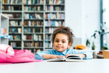 happy african american kid sitting and looking at camera near book and pink backpack