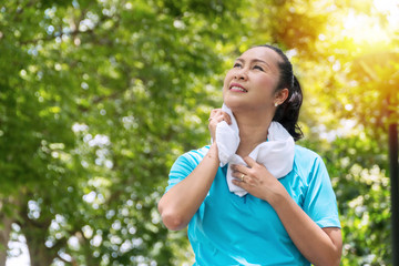 Elderly lifestyle concept. Attractive senior women 50s smiling while wiping her throat with white towel on neck after exercises in the public park.