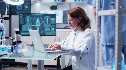In modern laboratory scientist woman types on laptop while her team is walking in the background