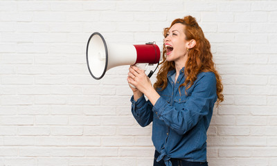 Redhead woman over white brick wall shouting through a megaphone