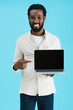 © LIGHTFIELD STUDIOS - smiling african american man pointing at laptop with blank screen isolated on blue