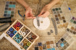 Top view of female hands making a macramé bracelet with kumihimo on a wooden table with tools, spools of thread, natural stones and colored beads
