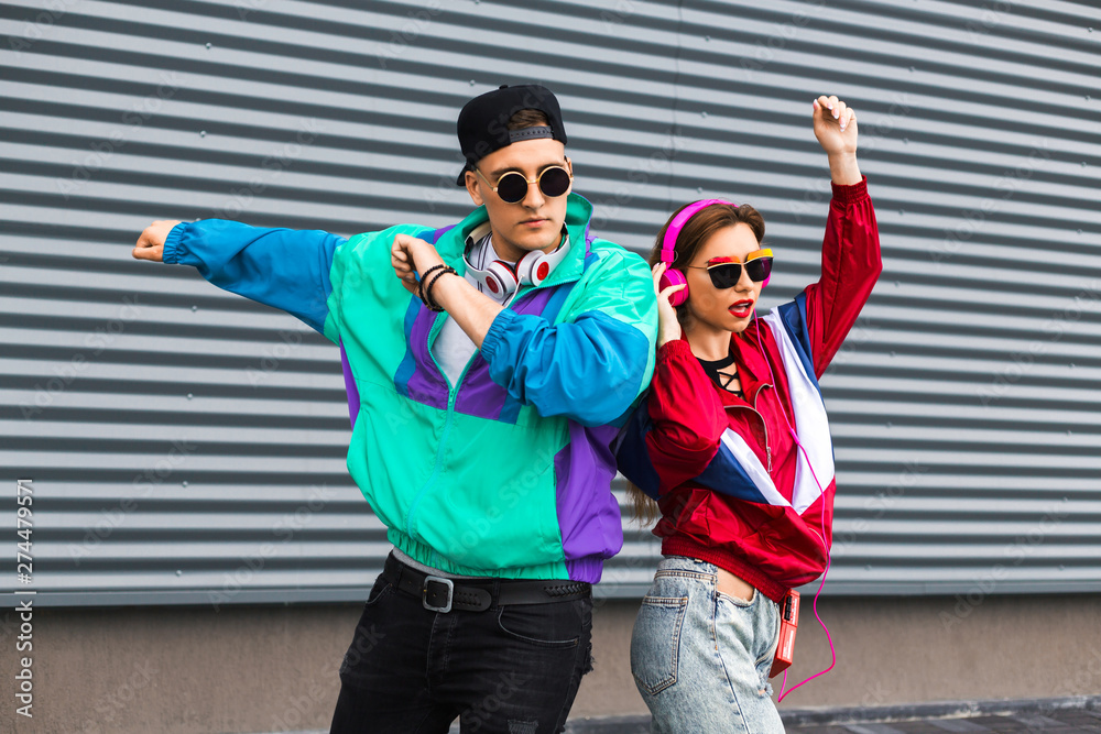 Back in time 90s 80s. Stylish young man in a retro jacket and a girl in red and with a vintage cassette player, against a steel wall, fashion trends, a street image - obrazy, fototapety, plakaty 