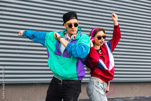 Fototapeta na szkło Back in time 90s 80s. Stylish young man in a retro jacket and a girl in red and with a vintage cassette player, against a steel wall, fashion trends, a street image