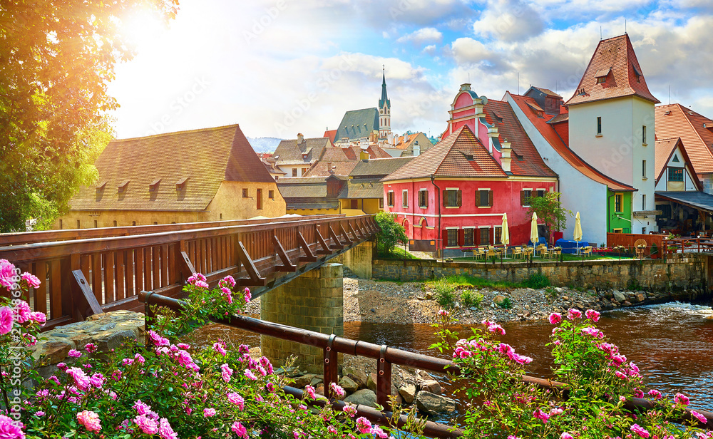 Czech Krumlov, (Cesky Krumlov), Czech Republic. Wooden bridge over river Vltava. Vintage picturesque old town with colorful houses and chapel of church. Rose flowers on bank. Sunny summer day. - obrazy, fototapety, plakaty 
