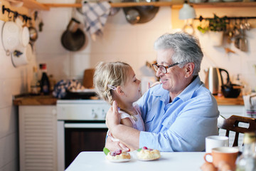 Wall Mural - Happy grandmother is hugging granddaughter in cozy home kitchen. Senior woman and cute little child girl are smiling. Kid is enjoying kindness, warm hands, care, support. Family is cooking together.