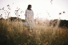 Stylish Girl In Rustic Dress Standing Among Wildflowers And Herbs In Sunny Meadow  In Mountains. Boho Woman Relaxing In Countryside At Sunset, Simple Life. Atmospheric Image. Space Text
