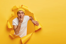 Image Of Frightened Puzzled Guy Covers Mouth, Stares With Scared Expression, Cannot Believe His Eyes, Poses In Ripped Paper Hole, Feels Embarrassed, Stands Against Yellow Background. Omg Concept