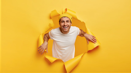 Horizontal shot of happy young man gazes something nice and awesome, dressed in casual outfit, keeps hands on edges of hole in yellow paper background, poses chill and carefree. Good mood concept