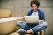 © iuricazac - Indoor shot of young man with curly hair using laptop for chatting online with friends, browsing Internet, sitting on the stairs at concrete wall. Candid male reading and typing on his laptop computer