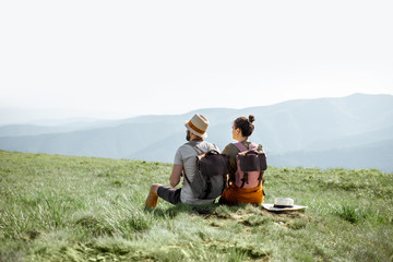 Young couple sitting on the green meadow, traveling with backpacks in the mountains during the summer time