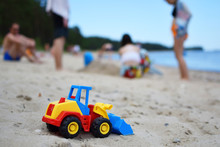 Toy Tractor Bulldozer On Beach Sand And Blurred Silhouette Of People And Sea On Background. Leisure Time With Friends And Family And Summertime Vacation Concept.