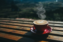 A Cup Of Hot Coffee Or Cappuccino Is Placed On An Outdoor Wooden Table In The Morning By The Smoke Rising From The Red Cup.