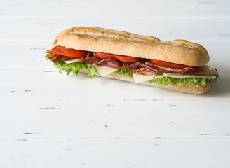 Fresh baguette sandwich with meat, sliced cheese, tomatoes and fresh lettuce on white wood background. copy space