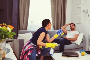 couple relationship concept, marriage  living problems and conflict, young couple in living room woman cleaning and vacuuming house, man sitting on sofa watching television, home jobs gender equality 