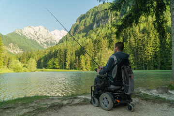 Wall Mural - young man in a wheelchair fishing at the beautiful lake on a sunny day, with mountains in the back