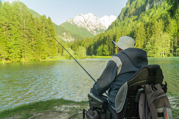 Wall Mural - young man in a wheelchair fishing at the beautiful lake on a sunny day, with mountains in the back