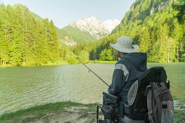 Wall Mural - young man in a wheelchair fishing at the beautiful lake on a sunny day, with mountains in the back