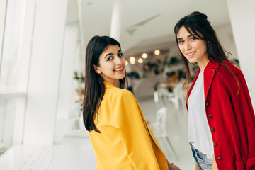 Wall Mural - Beautiful cheerful attractive young women look back and posing on camera together. Wear red and yellow jacket. Stand in white cafe room. Stylish models together.