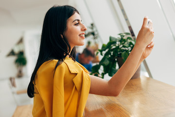 Wall Mural - Young stylish turkish modern buisnesswoman taking picture with phone camera. Sitting in white coworking room alone and work. Wear yellow casual jacket. Sit at table.