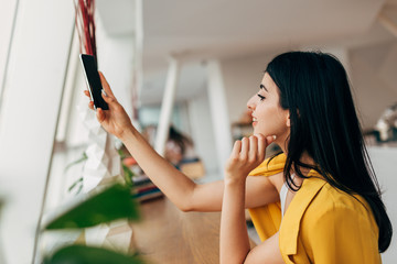 Wall Mural - Attractive dark-haired young woman taking picture with phone camera. Work in coworking white room. Partner alone. Woman wear yellow casual jacket.