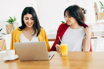 Wall Mural - Beautiful attractive young women in coworking space. Working together. Stylish businesswomen in cafe. Model on left typing on keyboard. Woman on right drink orange juice. Sit on table.