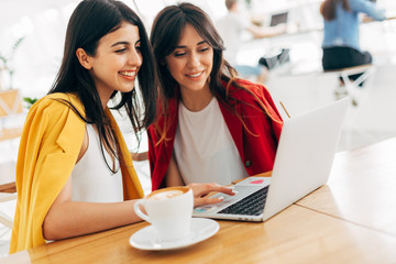 Wall Mural - Two young businesswoman work together in white room. Look at laptop and smile. Business partners in coworking space. Stylish models in red and yellow jacket. Coffee cup on table.