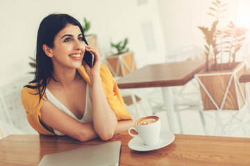 Wall Mural - Nice cheerful attractive young woman sitting at table and talking on phone. Smiling. Cup of coffee and laptop on table. Room with white walls or coworking.