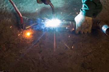 A strong man is a welder in a black T-shirt, in a welding mask and welders leathers, a metal product is welded with a welding machine in the garage, blue sparks fly to the sides