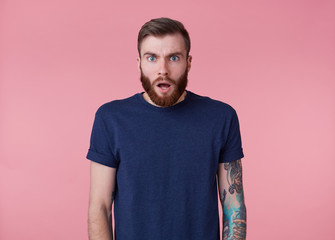 Wall Mural - Photo of young handsome amazed red bearded manman in blank t-shirt, hears unbelievable news, looks surprised, stands over pink background with wide open mouth and eyes.