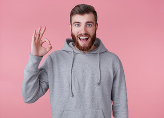 Wall Mural - Look at this cool guy! Young handsome positive red bearded man in gray hoodie, looks happy and broadly smiles, stands over pink background and shows okay gesture.