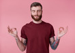 Portrait of young attractive red bearded man in blank t-shirt, looks peaceful and calm, smiles, stands over pink background with closed eyes and showing om gesture.